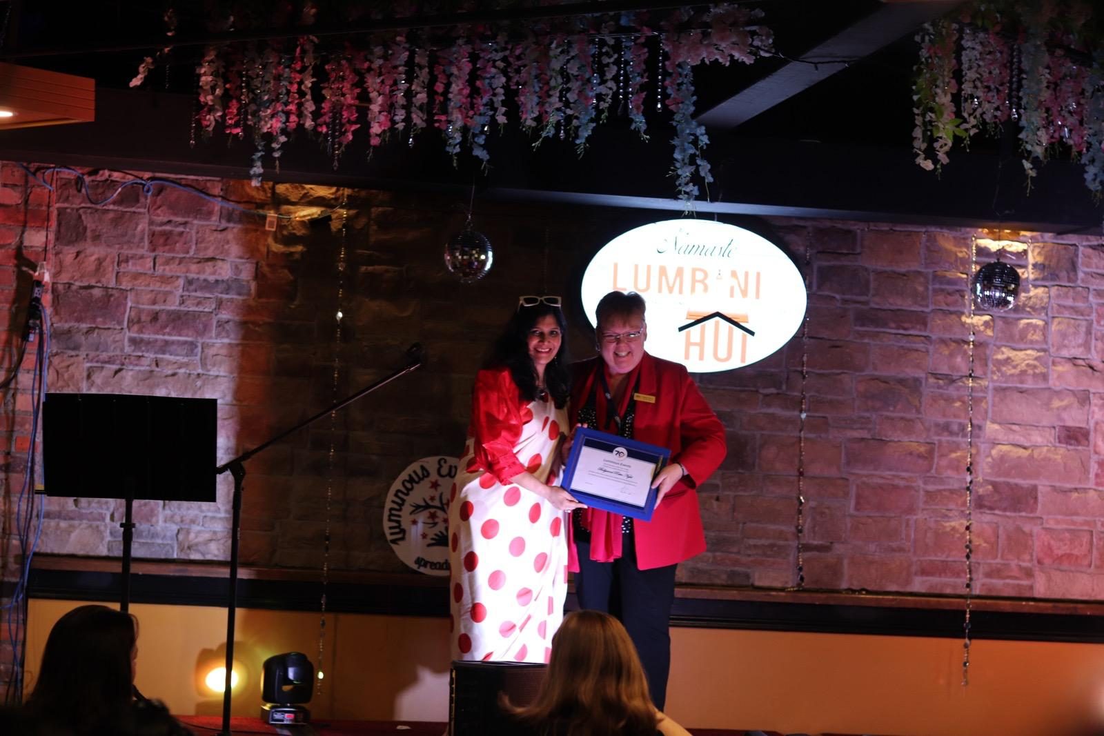 A woman in a red and white polka-dot saree and a guest in a red blazer holding a framed certificate for Bollywood Retro Night on stage
