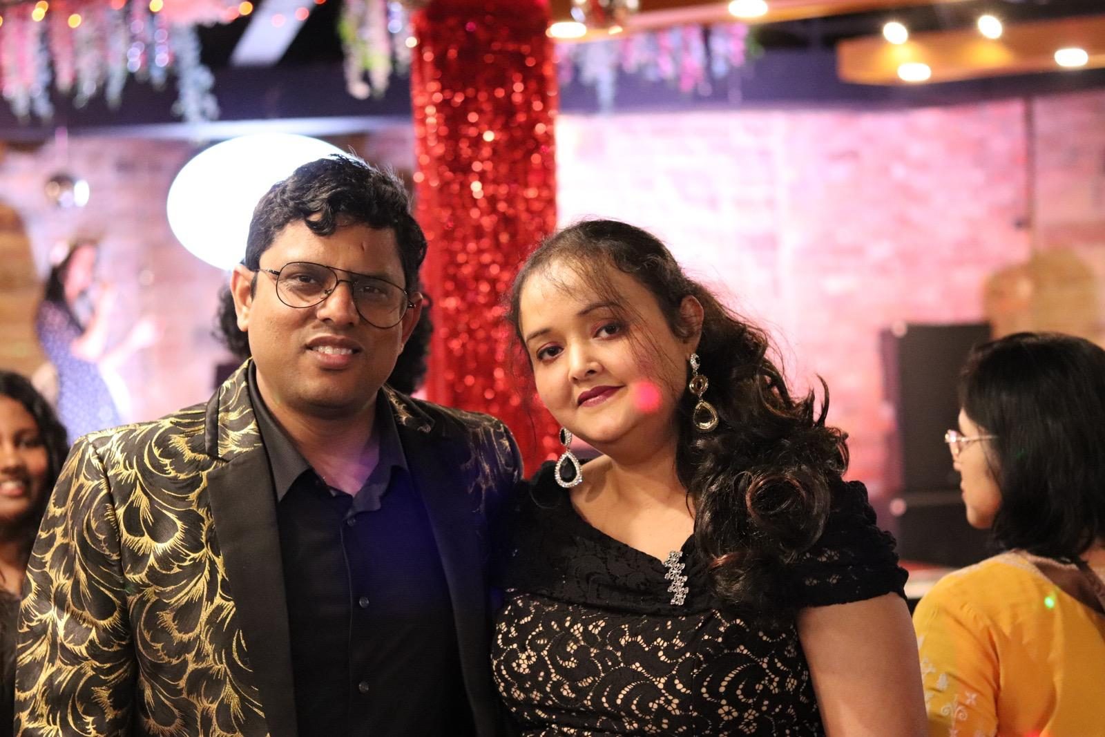 A couple in a gold-patterned blazer and a black lace dress posing in front of a red sequinned pillar