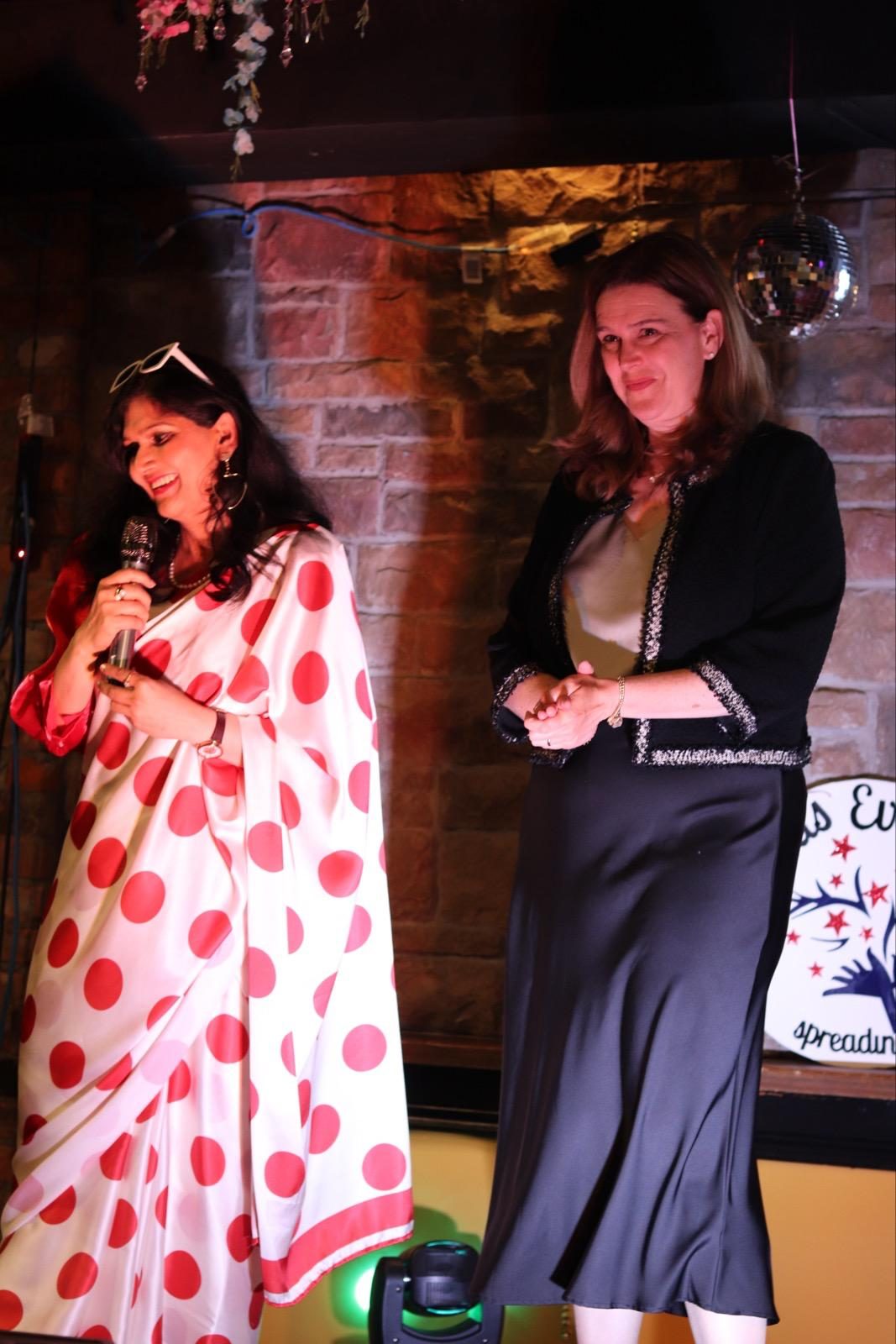 Two guests on stage, one in a red and white polka-dot saree speaking into a microphone