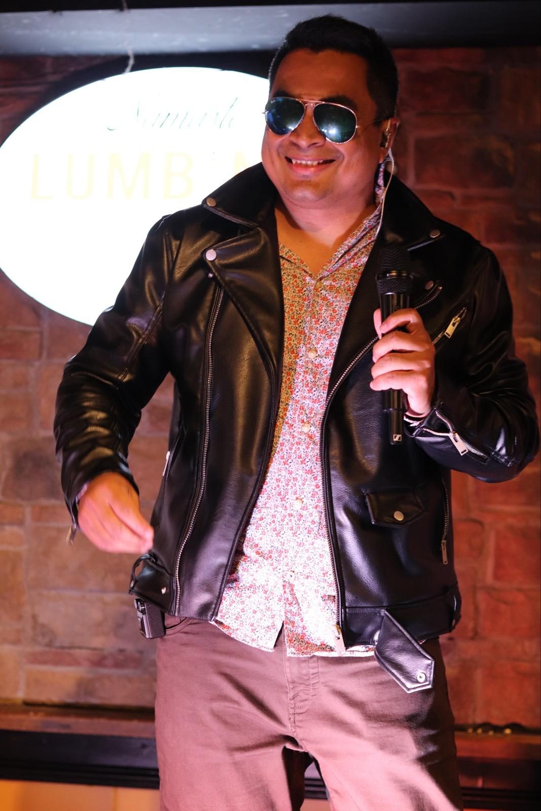 A smiling singer in a leather jacket, floral shirt and mirrored sunglasses holding a microphone