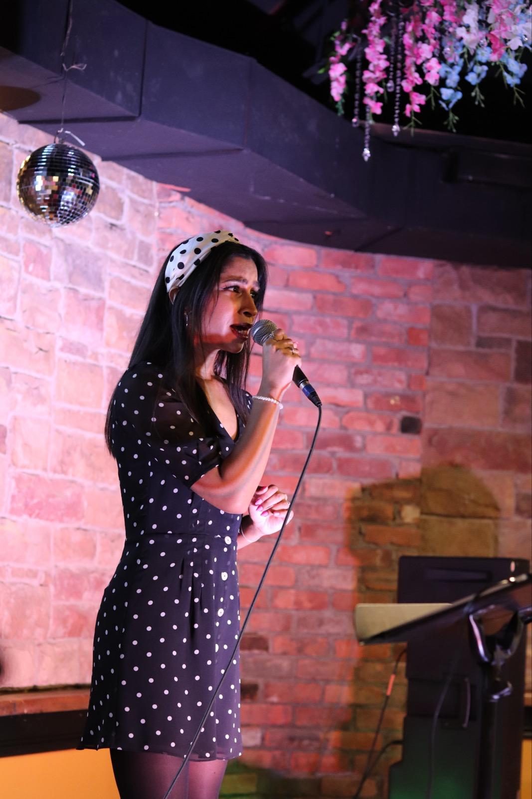 A singer in a polka-dot dress and headband performing under hanging flowers and a disco ball