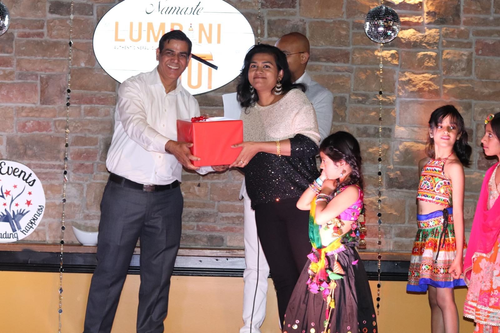 A prize being presented on stage in front of the Lumbini Hut sign, with young guests in colourful lehengas looking on