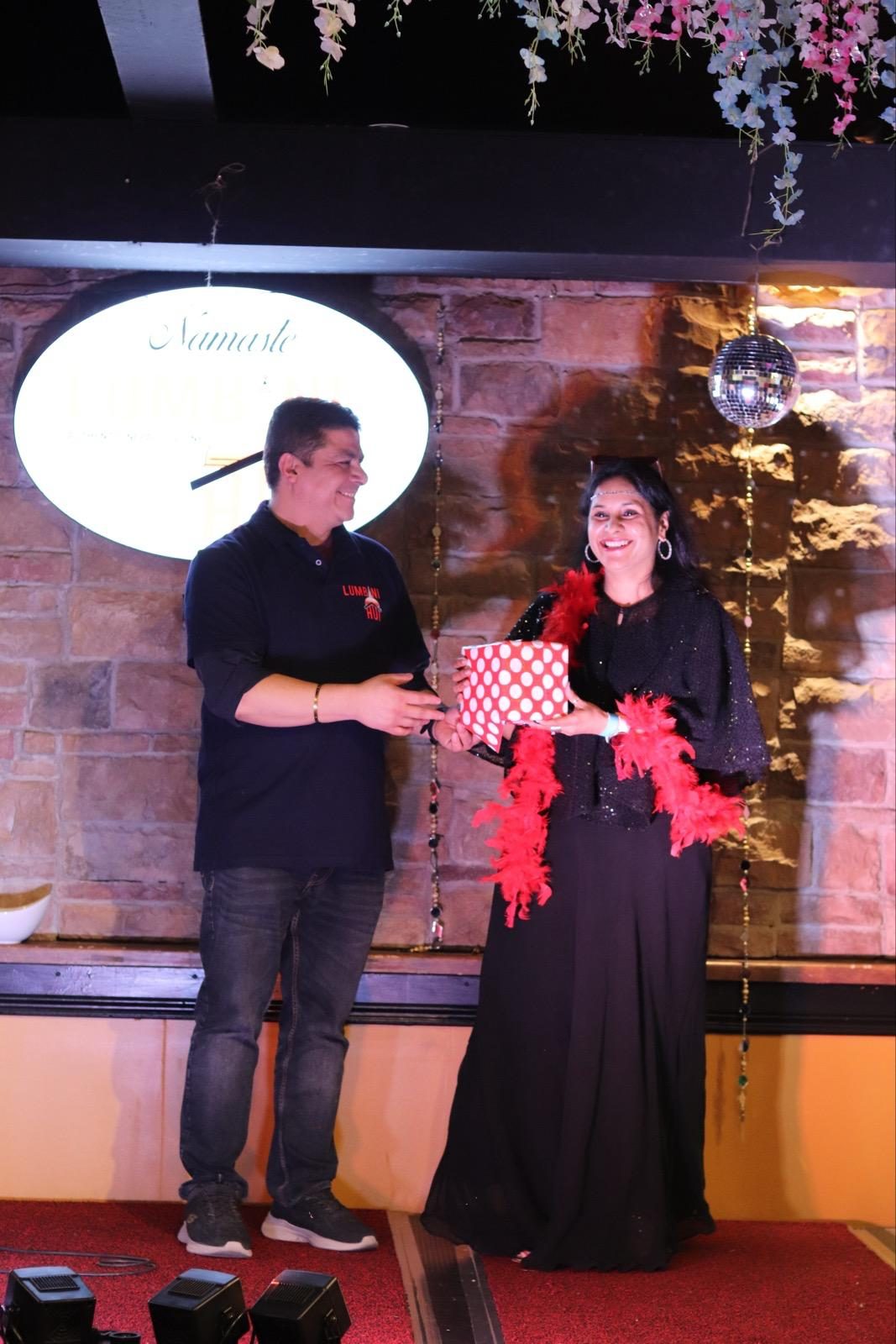 A guest in a black sequinned outfit and red feather boa receiving a polka-dot gift box from a member of the Lumbini Hut team