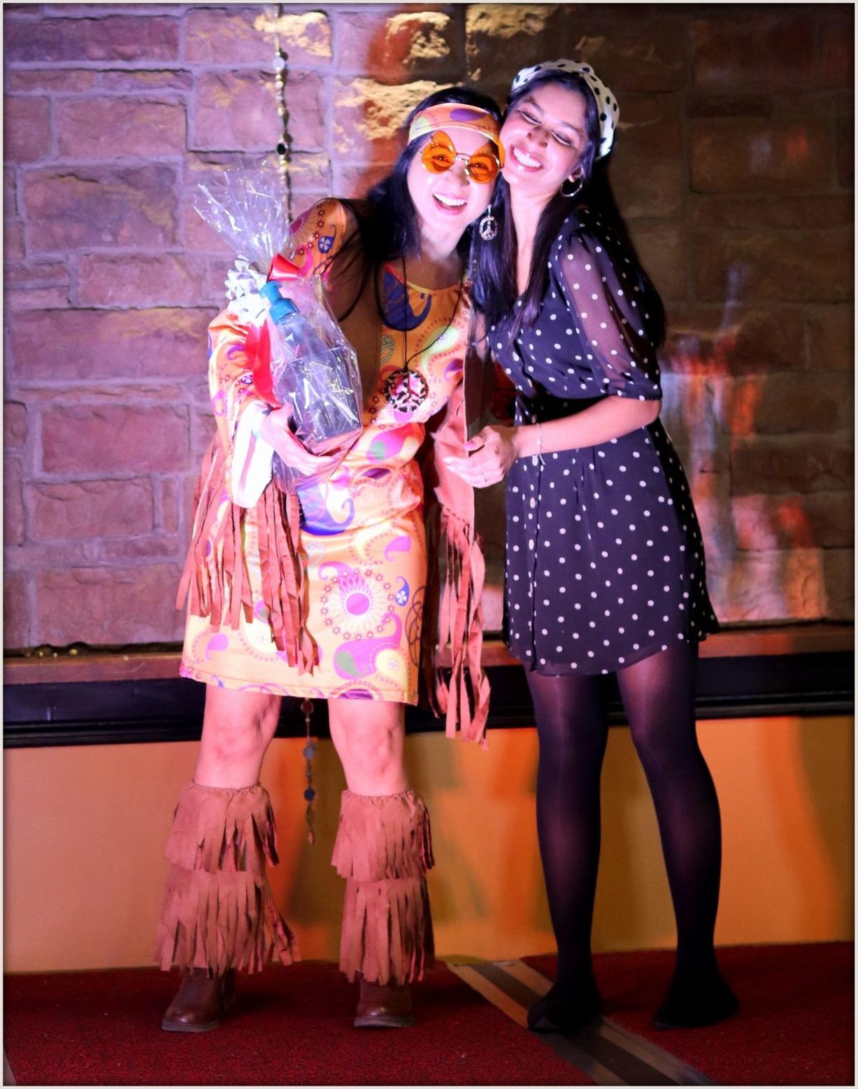A winner in an orange paisley hippie dress with fringe boots, holding a gift basket and laughing as she is hugged by a guest in a polka-dot dress