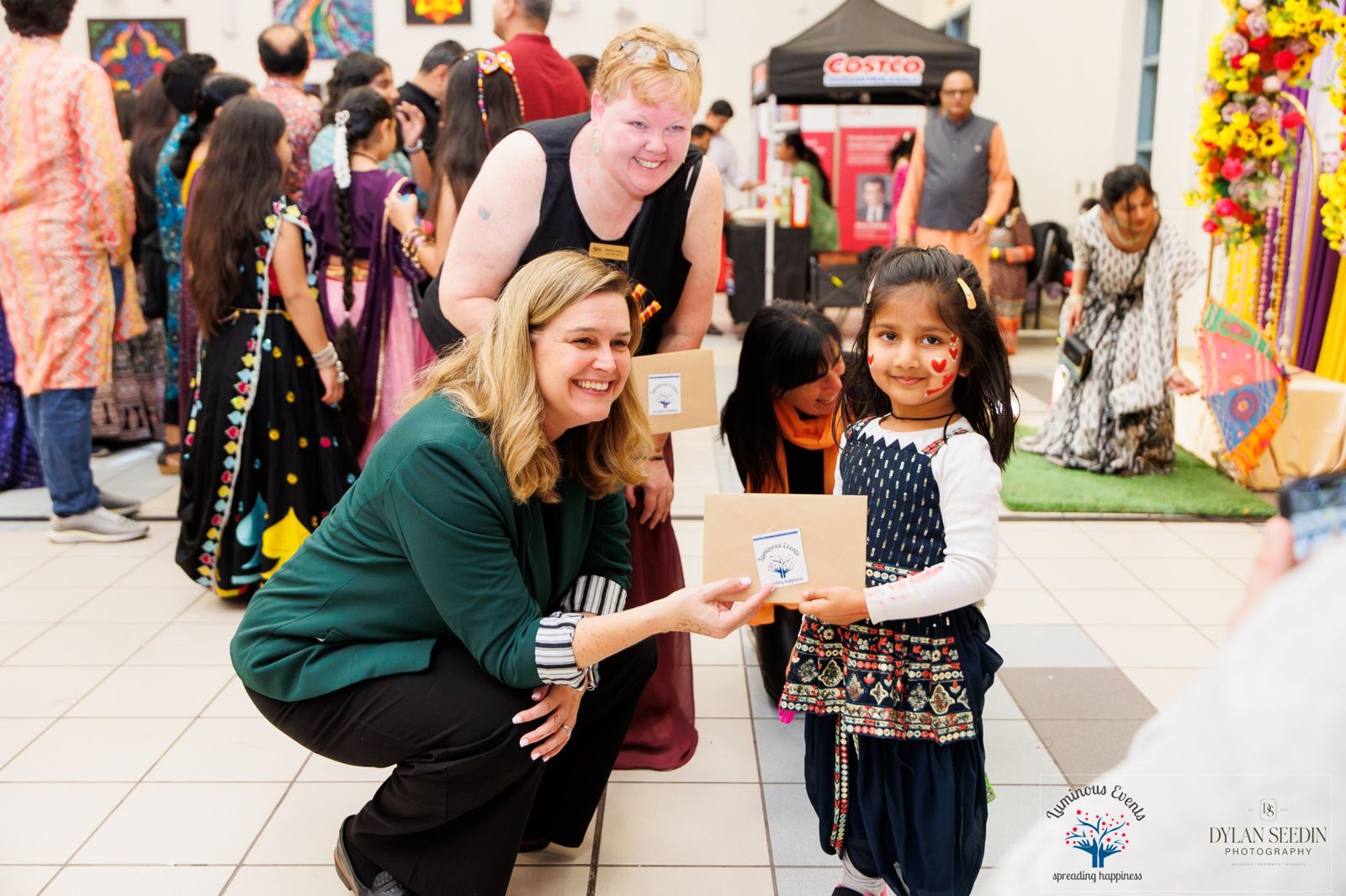 Guests of honour crouching to hand a Luminous Events gift envelope to a young girl with heart face paint