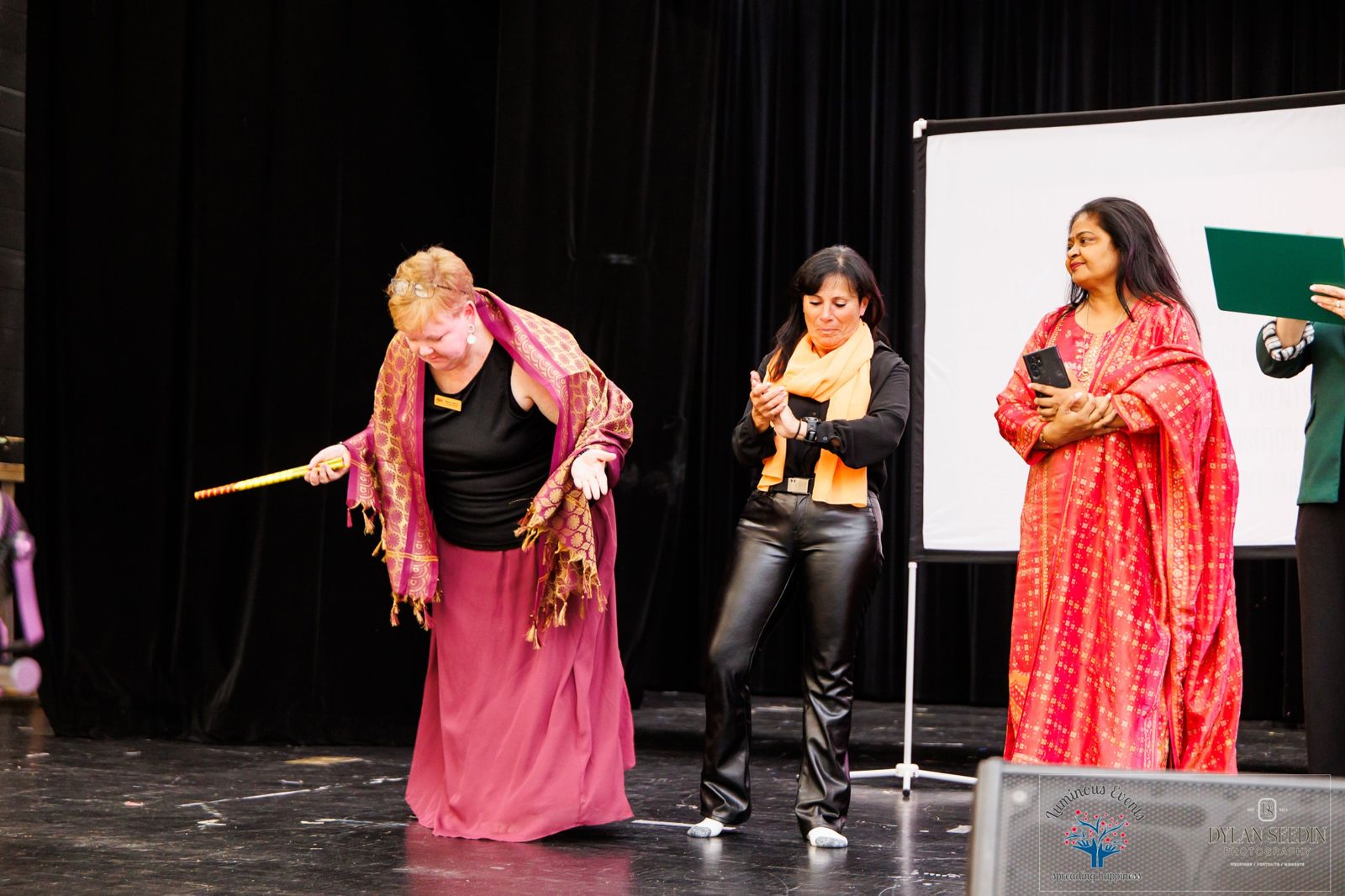 A guest on stage with a dandiya stick and shawl taking a joyful bow while organizers applaud