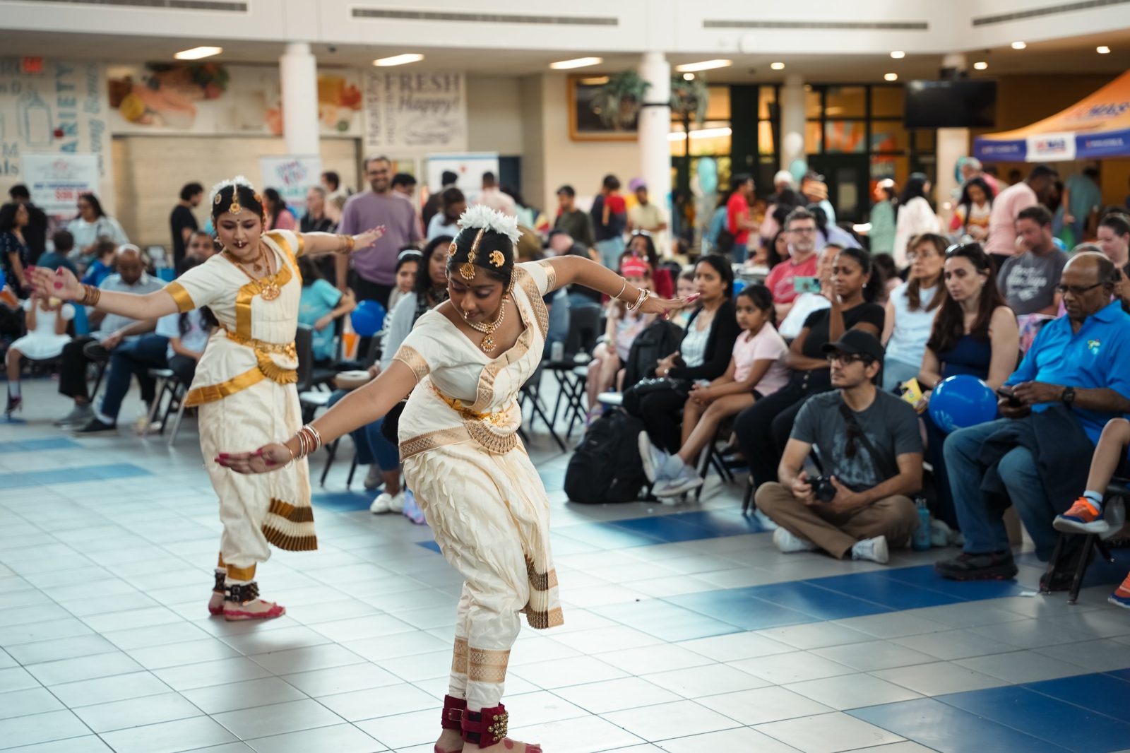 Two classical dancers in cream and gold costumes performing on the tiled floor in front of seated guests
