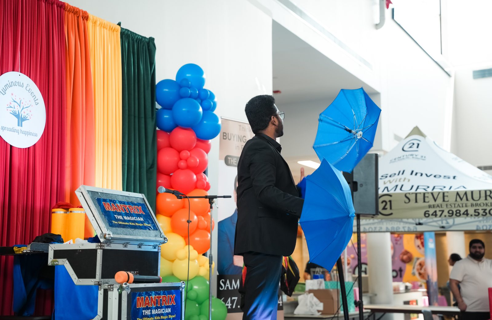 Mantrix the Magician on stage with blue umbrellas beside his magic case and the Luminous Events backdrop