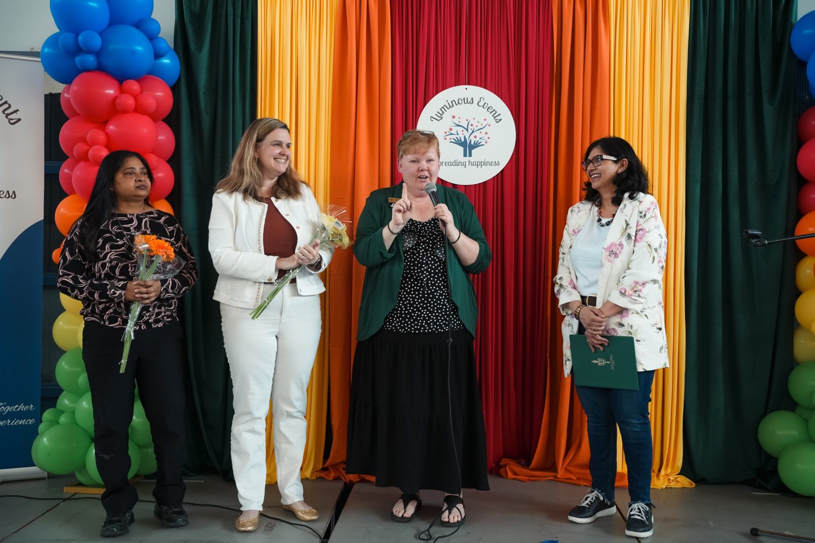 Four honoured guests on stage in front of the colourful curtain backdrop, two holding flowers
